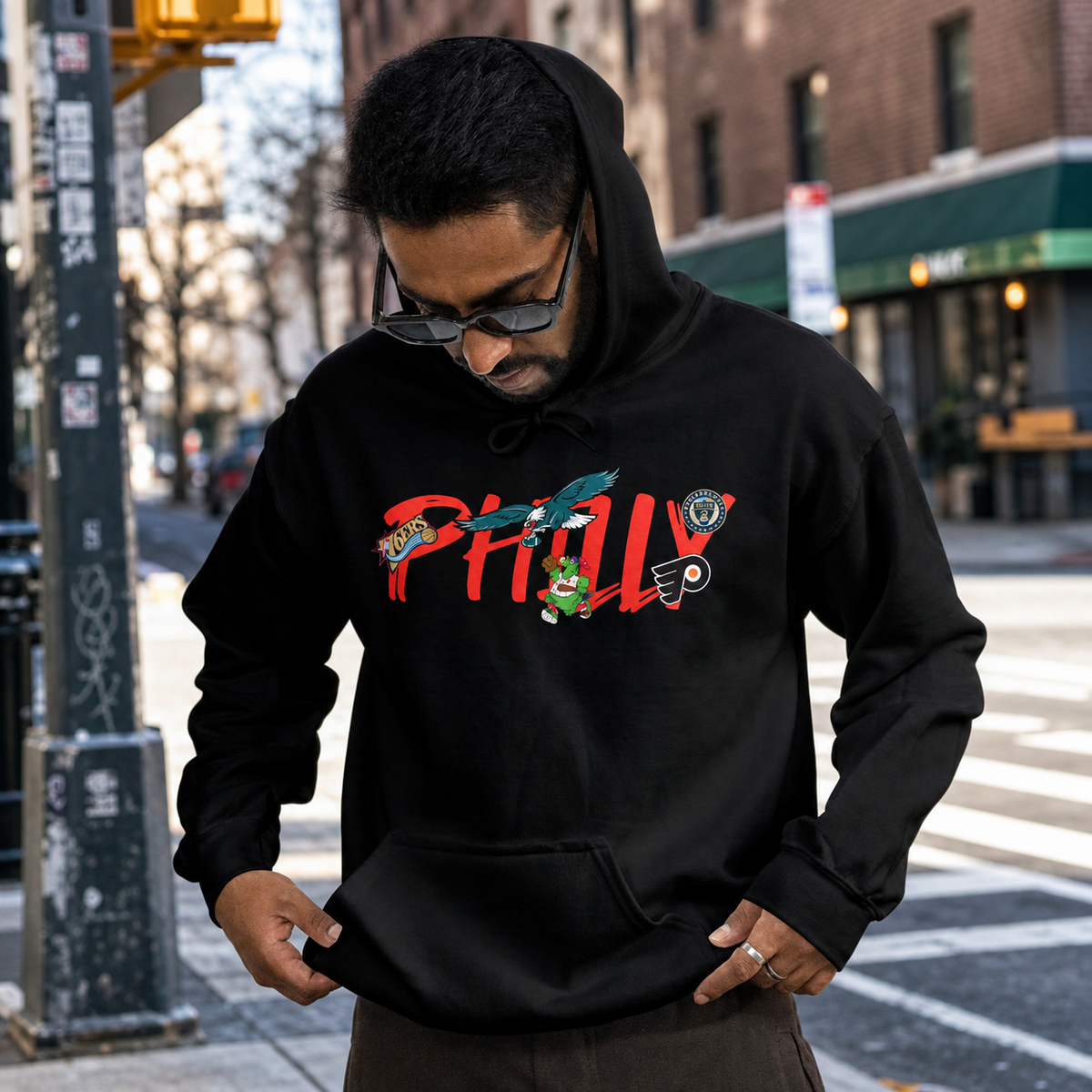A man wearing a black hoodie with a colorful 'PHILLY' graphic featuring cartoon characters, standing on a city street.